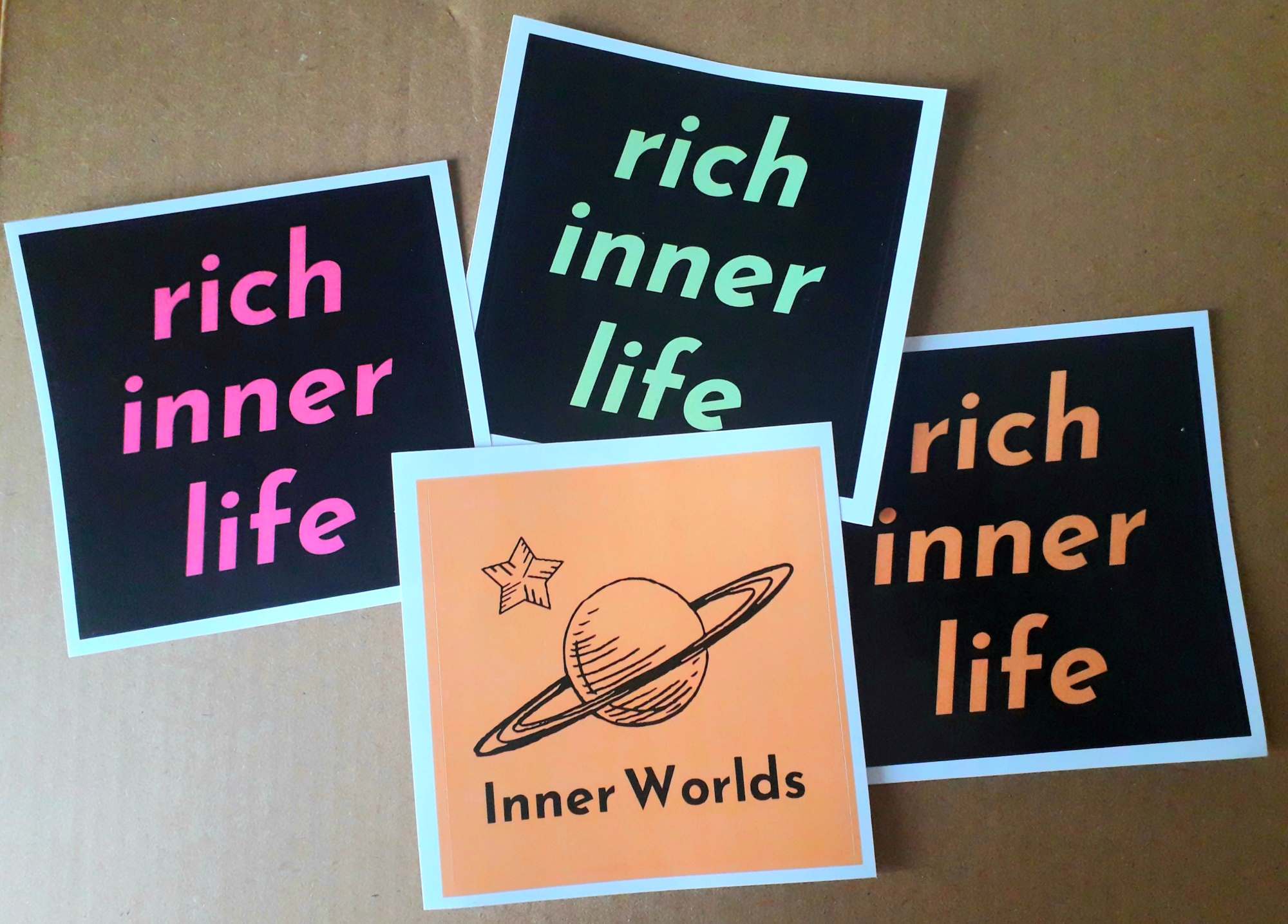 Large square stickers, one is light orange with the Inner Worlds logo and the others are black with neon text reading "rich inner life"